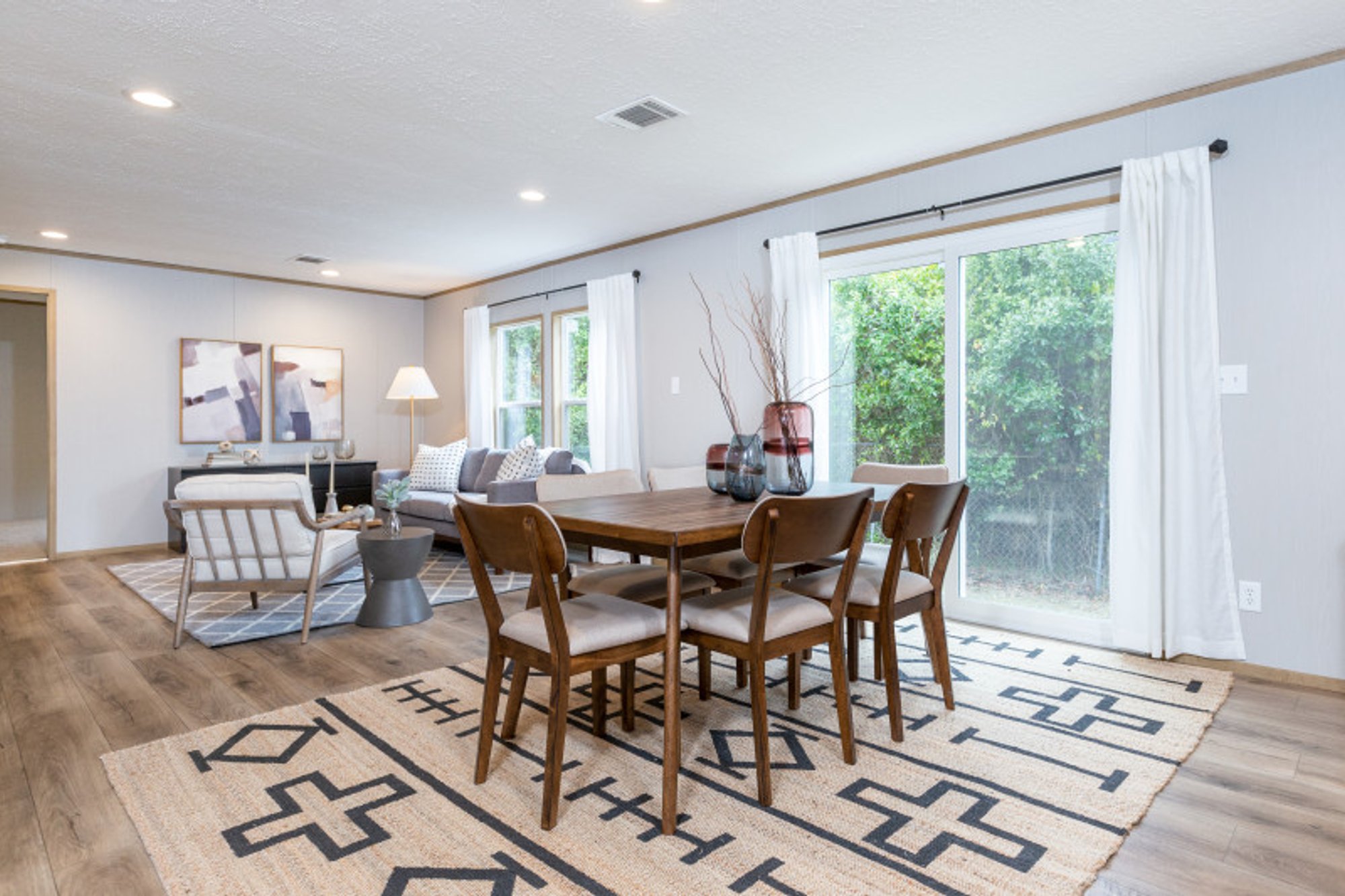 Open-concept dining and living area with light wood flooring, mid-century modern furniture, large windows with white curtains, and natural light.