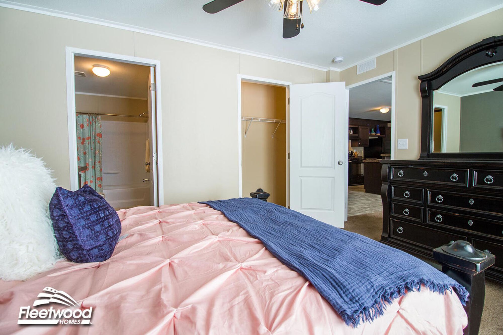 Bedroom with a pink comforter and navy throw blanket on the bed, black dresser with mirror, ceiling fan, and open doors leading to a walk-in closet, bathroom with shower-tub combo, and view into the kitchen.