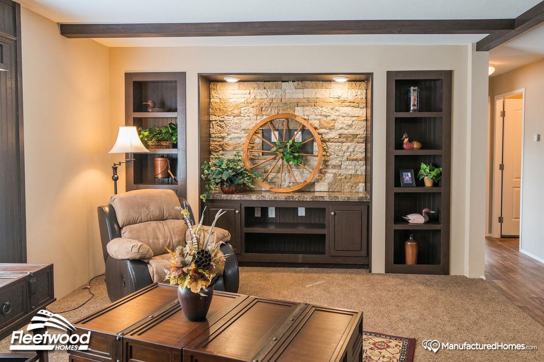 Cozy living room in a manufactured home, featuring beige recliner seating, a dark wood coffee table, and decorative plants on plush carpet with a patterned area rug.