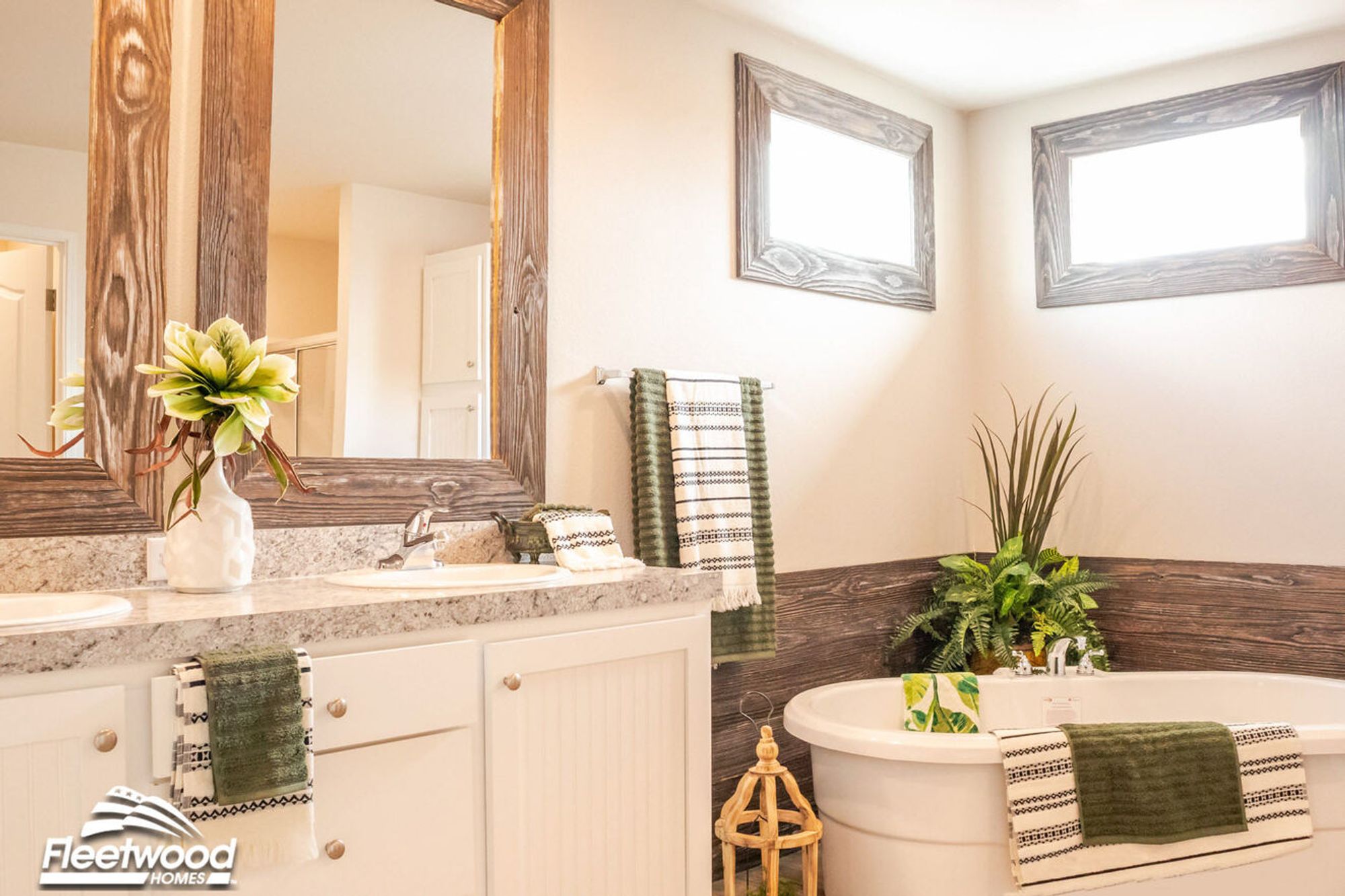 Bathroom with double sinks, granite countertops, wood-framed mirrors, and a soaking tub in the corner surrounded by wood and greenery décor.