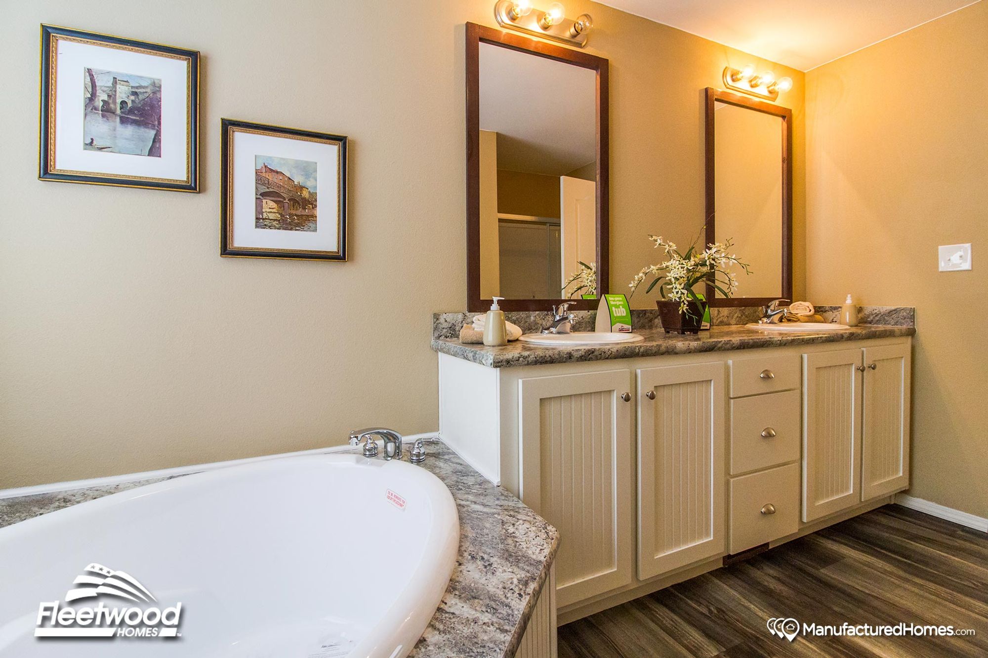 Inviting bathroom with a large corner soaking tub, dual-sink vanity with beadboard cabinets, granite countertops, and two framed mirrors under warm lighting.
