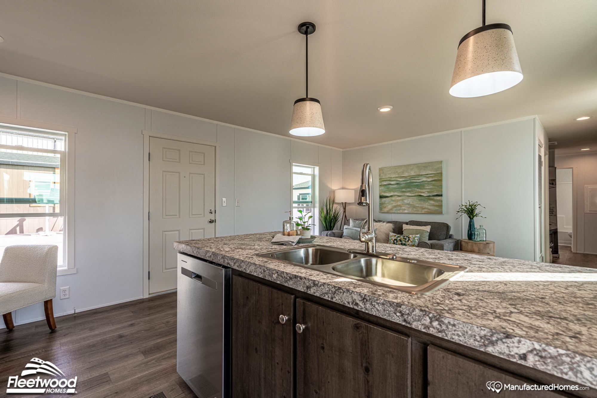 Open-concept kitchen and living space featuring a marbled countertop island with a stainless steel sink, pendant lighting, and a cozy sitting area with coastal-themed decor.