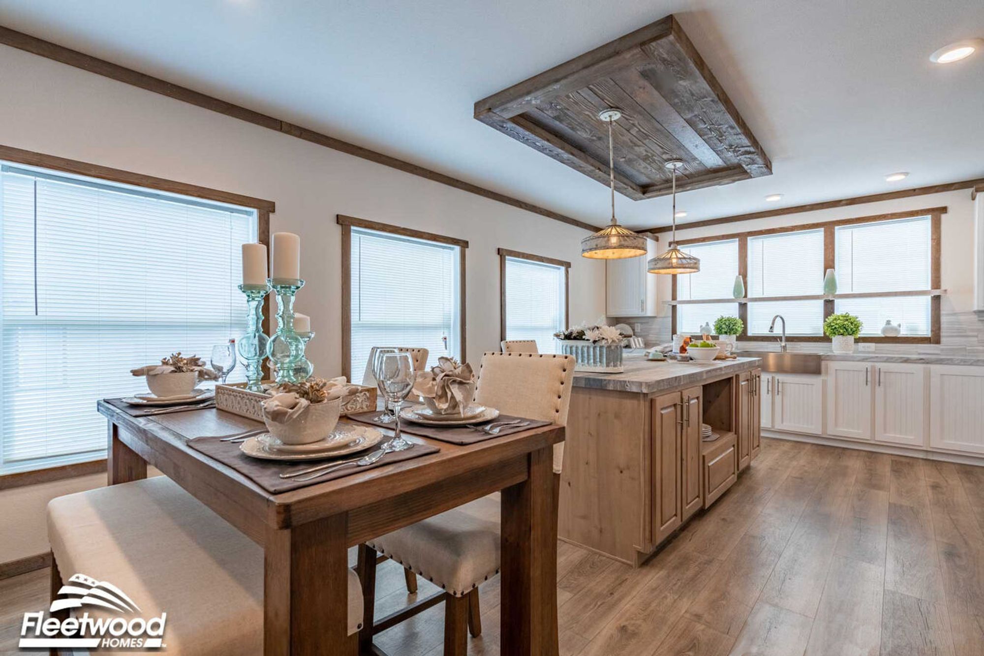 Dining area with a wooden table set for four, next to a rustic kitchen featuring a large island, pendant lighting, white cabinetry, and a farmhouse sink under three windows.
