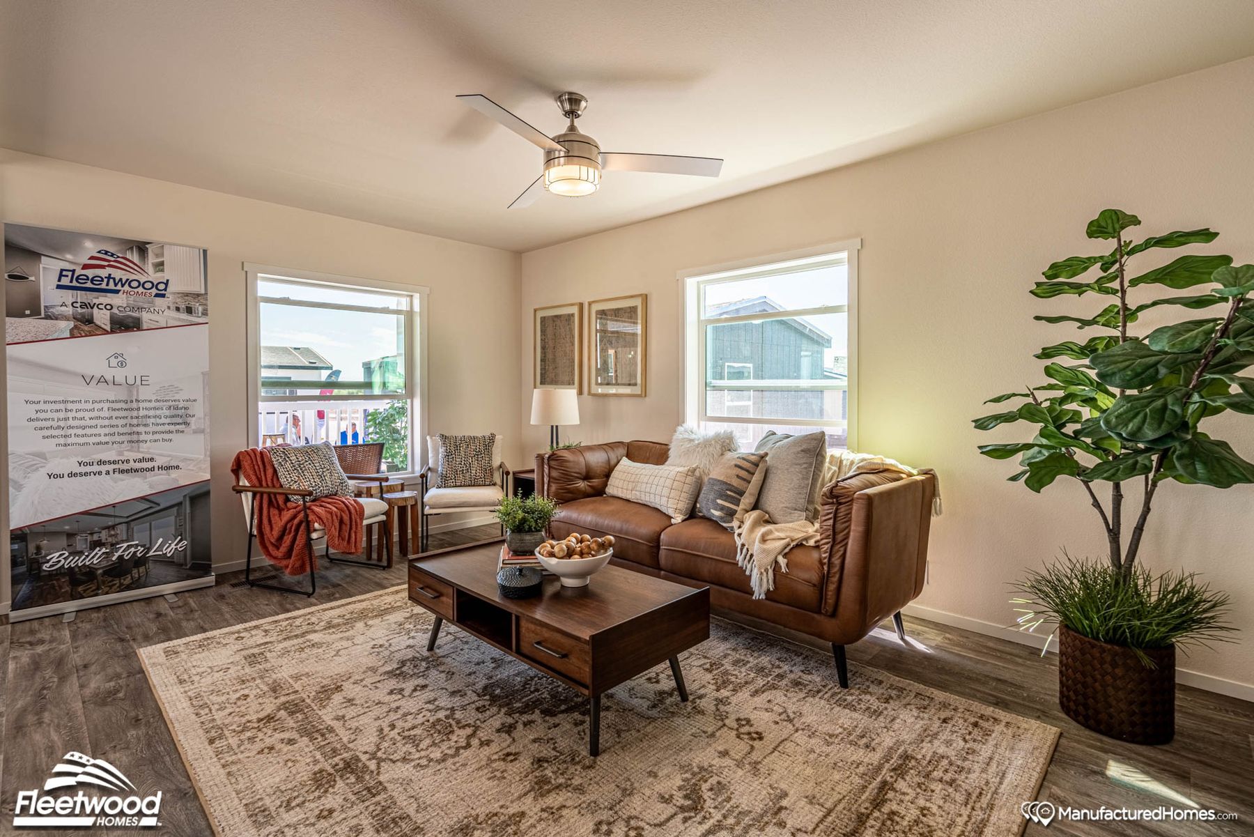 Cozy living room with a brown leather sofa, two accent chairs, a wood coffee table, and large windows. Decor includes neutral pillows, throws, a patterned rug, indoor plants, and a ceiling fan. A Cavco Idaho banner stands in the corner.