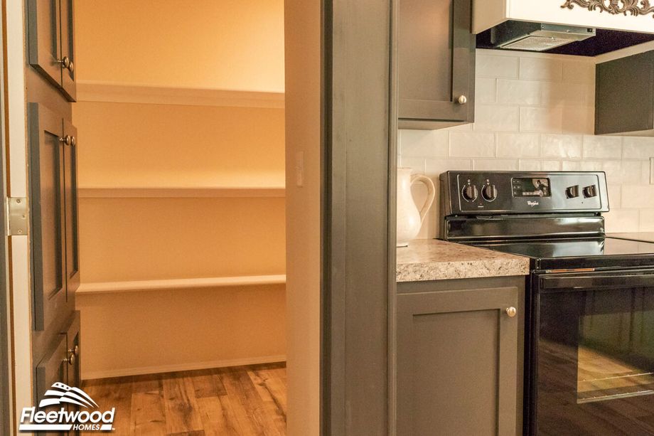Walk-in kitchen pantry with white built-in shelving, located adjacent to a black stove and granite-look countertops in a modern kitchen.