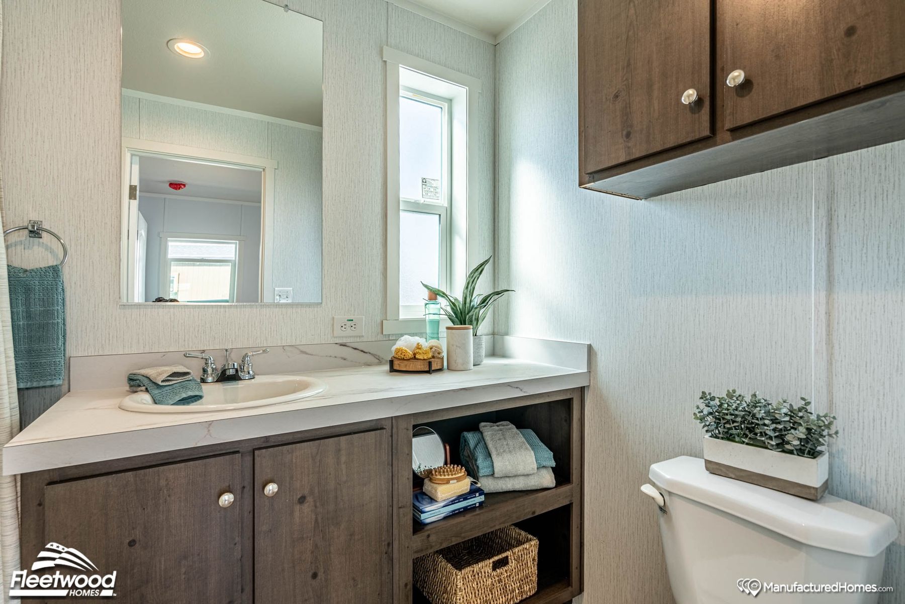 Bright and neatly organized bathroom featuring a single-sink vanity with marble-look countertop, wood cabinetry with open shelving, wall-mounted cabinet, and decorative elements like towels, plants, and baskets near a frosted vertical window.