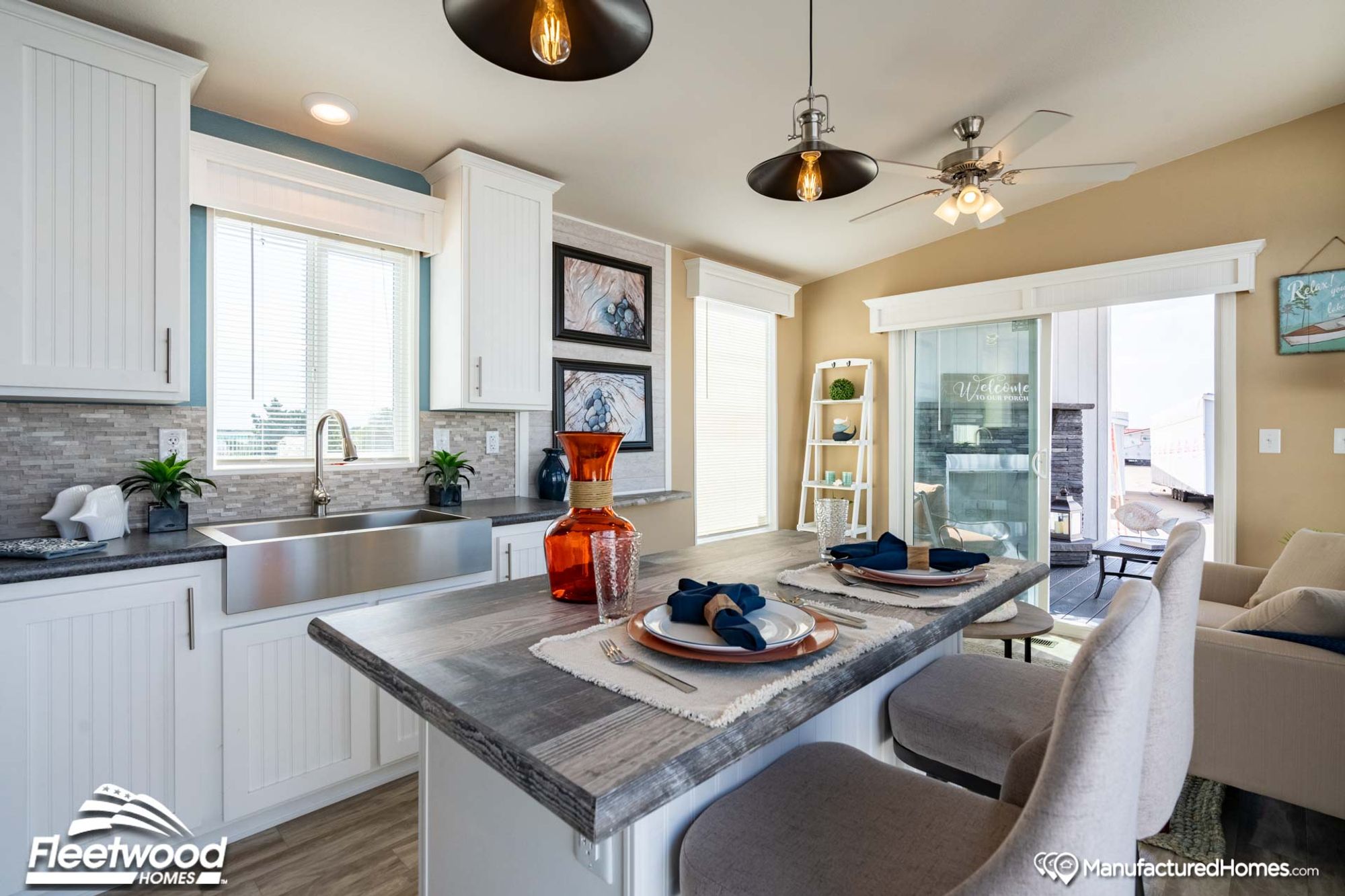 Stylish kitchen and dining area featuring a gray wood-look breakfast bar set with plates and navy napkins, white shaker cabinets, stainless steel farmhouse sink, pendant lighting, and sliding glass doors opening to a patio.