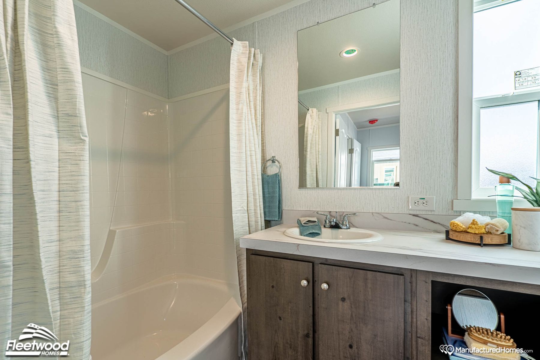 Modern bathroom with a curved shower-tub combo and curtain, single-sink vanity with marble-style countertop and dark wood cabinetry, large mirror, and decorative accents like towels, a plant, and a small vanity mirror near a frosted window.