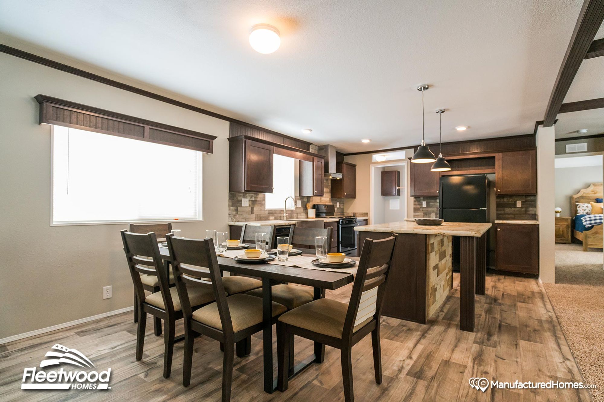 Stylish kitchen and dining area in a manufactured home featuring a wood-accented island, dark cabinetry, tile backsplash, and a six-seat dining table set for a meal.