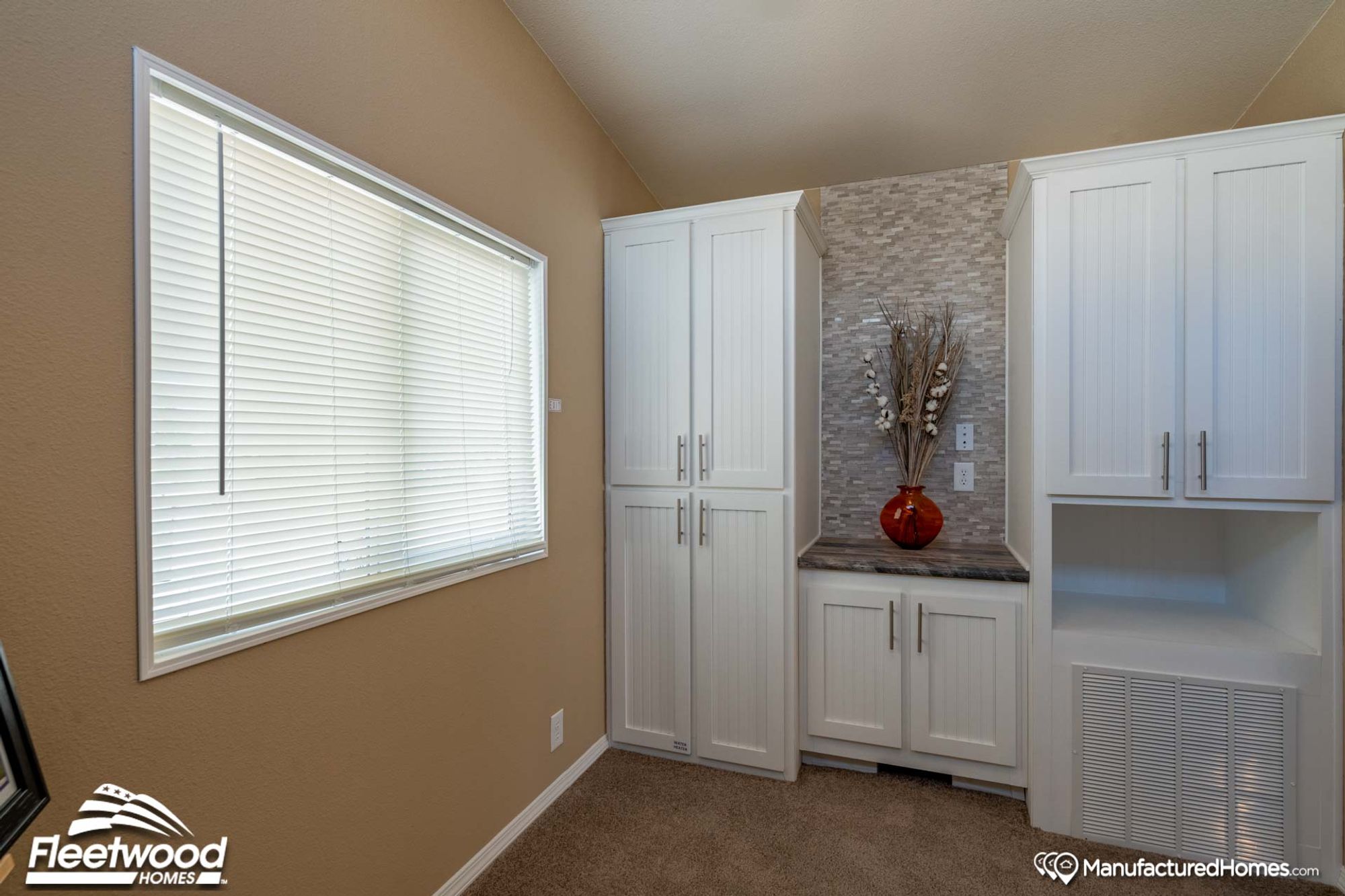 Compact utility or storage area with white shaker-style cabinets, stone backsplash accent, and a built-in niche decorated with a red vase and dried botanicals. A large window with blinds brightens the space.