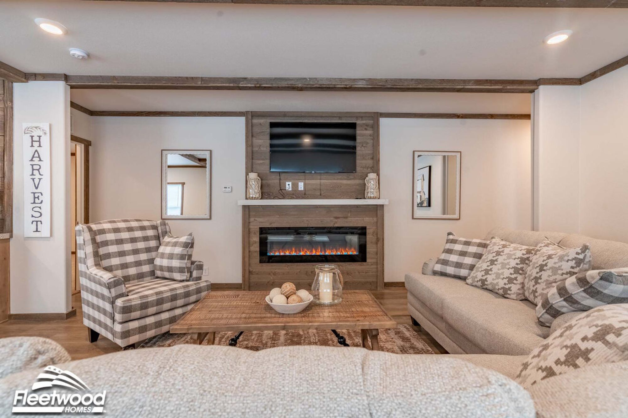 Cozy living room with wood-beam ceiling accents, neutral-toned sofas with patterned pillows, a plaid armchair, a rustic coffee table, and a wood-paneled fireplace wall with a mounted TV.