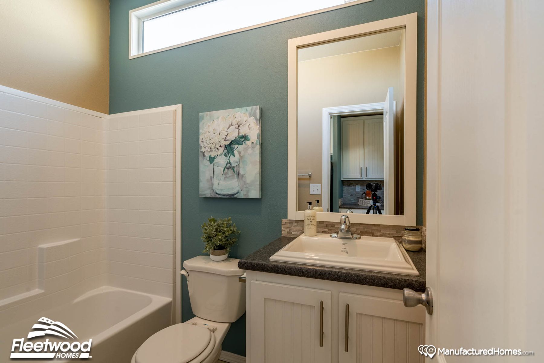 Bathroom with a white bathtub and shower combo, white vanity with dark countertop, and a rectangular framed mirror. A toilet sits below a small decorative plant, and the teal accent wall features a floral painting. A high horizontal window lets in natural light.