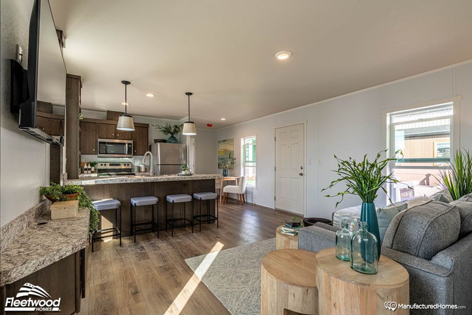 Open-concept living space with a view of the kitchen featuring a marbled island with barstools, pendant lighting, stainless steel appliances, and a cozy living area with wood-accented decor and greenery.