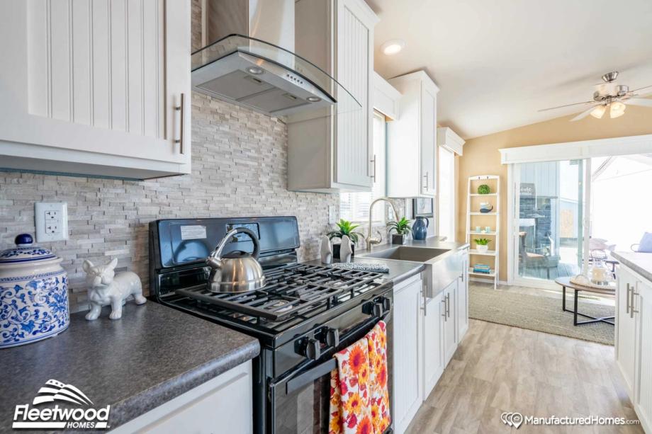 Modern kitchen with white shaker cabinets, stone backsplash, stainless steel farmhouse sink, and black Whirlpool gas stove with a floral towel hanging on the oven door. A glass range hood is mounted above the stove, and sliding glass doors lead to a patio area in the background.