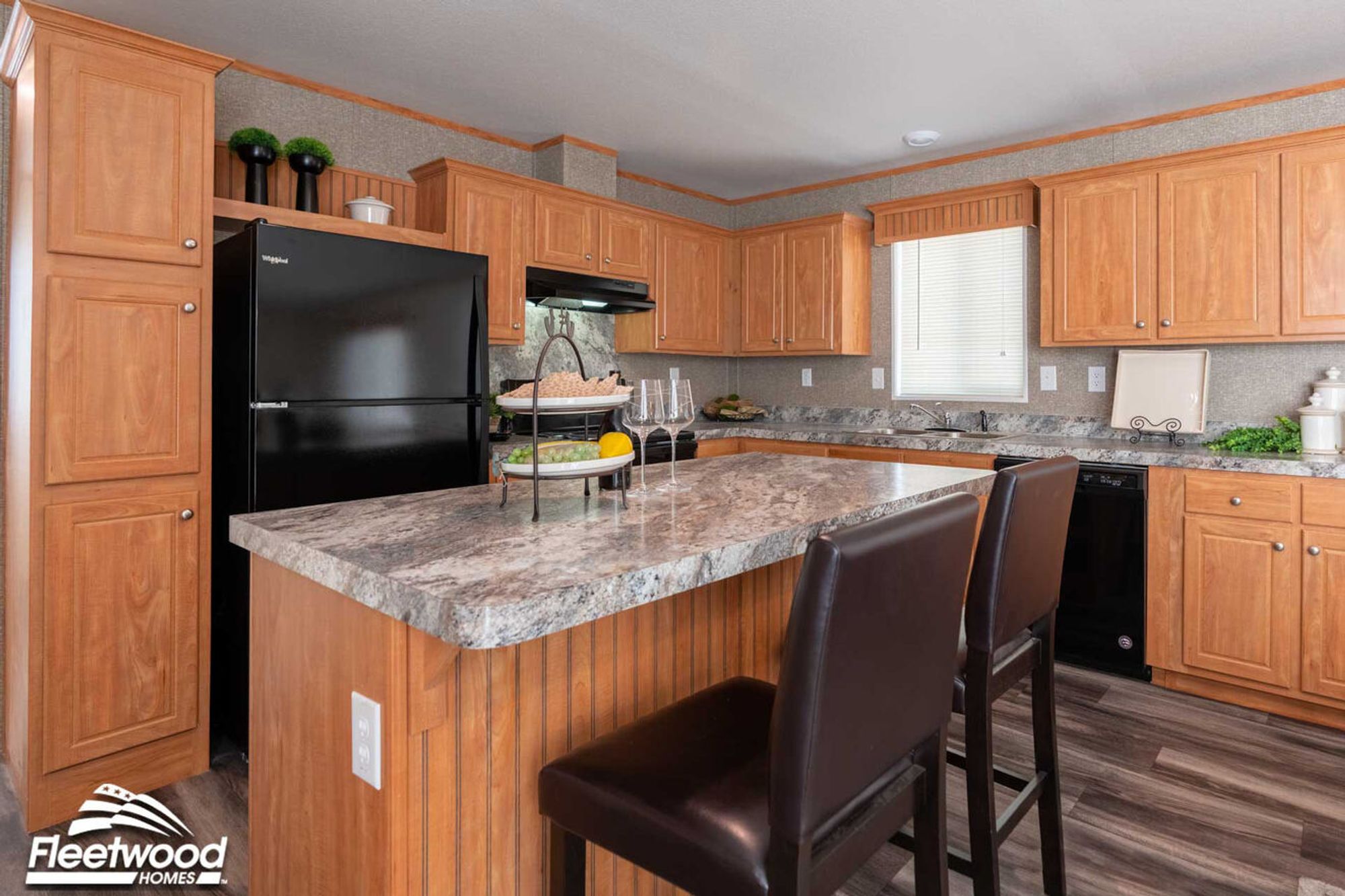 Kitchen with wood cabinetry, a large marble-look island with seating, black appliances including a refrigerator, stove, and dishwasher, and a window above the sink.