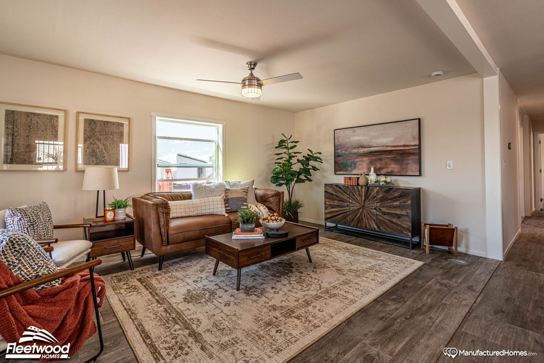 Cozy living room with a brown leather sofa, two accent chairs, a wood coffee table, and large windows. Decor includes neutral pillows, throws, a patterned rug, indoor plants, and a ceiling fan. A Cavco Idaho banner stands in the corner.