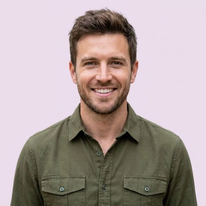 A smiling man with short brown hair and a beard, wearing a green shirt, against a light pink background.