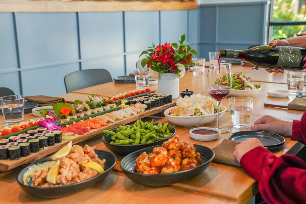 A table of sushi boards and various food dishes, with a person pouring wine into a glass.