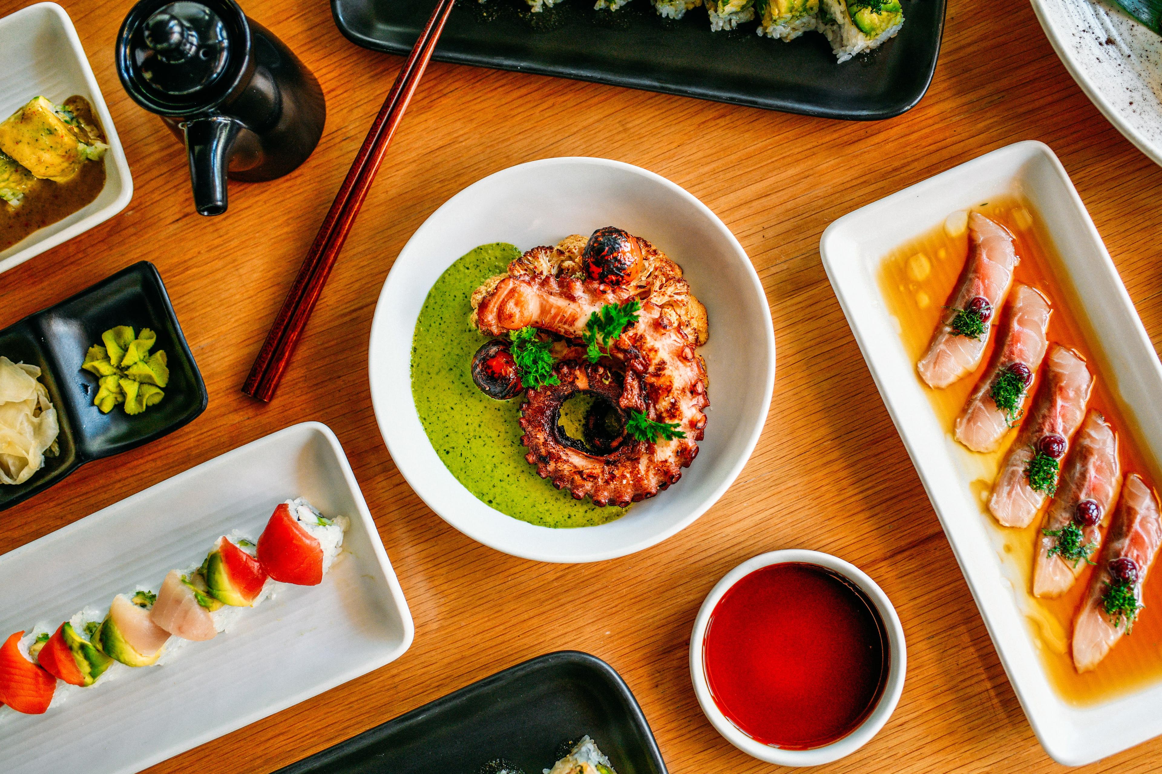 Overhead shot of grilled octopus, sashimi, and sushi dishes on a table.