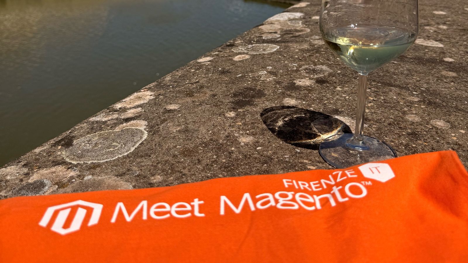 Orange Meet Magento Firenze banner on the stone wall of the Lungarno in Florence, with the Arno behind it, during the lunch break at Meet Magento Italy 2026.