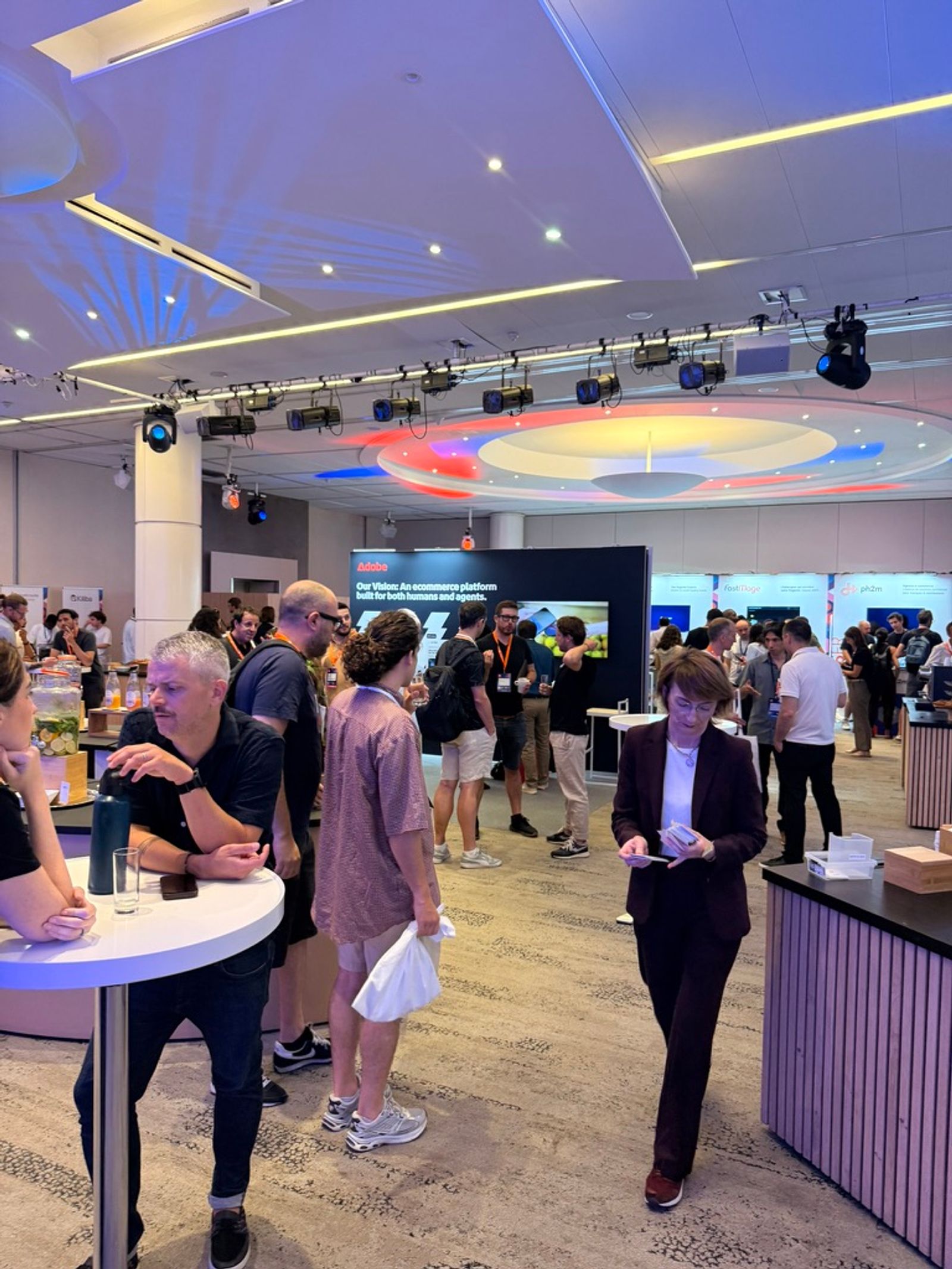 Meet Magento France 2026 venue interior at Les Salons de l'Aveyron in Paris, with attendees mingling around the Adobe Commerce booth and other sponsor stands.