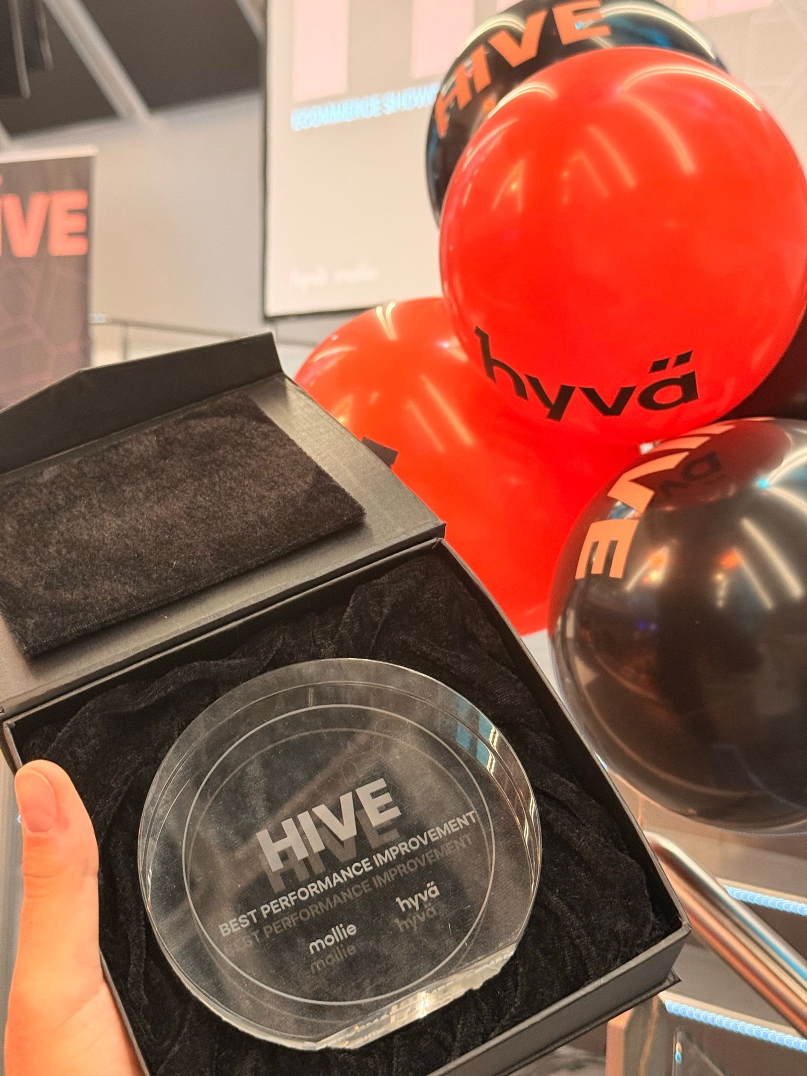Premiul HIVE Germany 2026 pentru Best Performance Improvement, un disc de cristal gravat cu siglele Mollie și Hyvä, în cutia lui, lângă baloane Hyvä.
