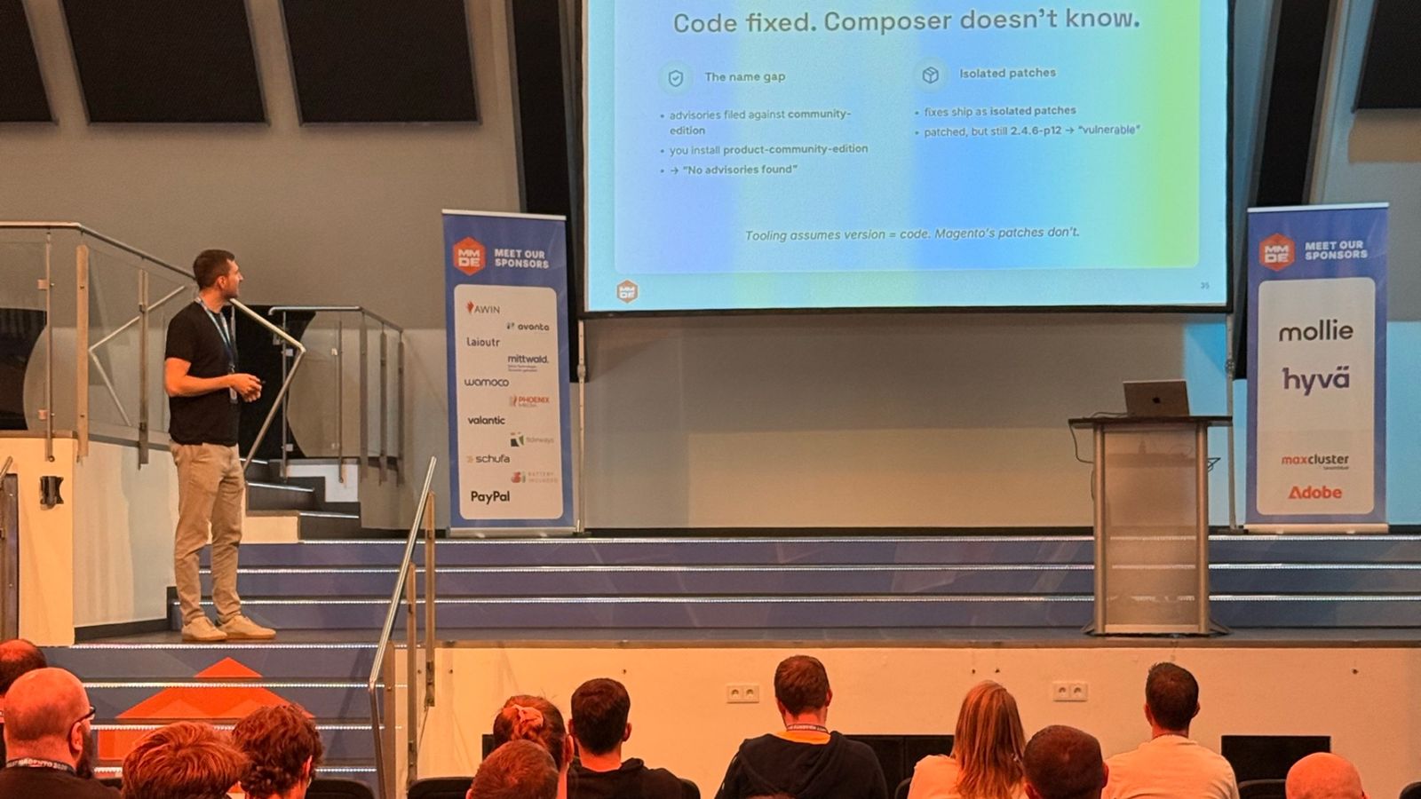Un vorbitor pe scena mare de la Meet Magento Germany 2026, lângă slide-ul „Code fixed. Composer doesn't know.”, despre diferența de nume și patch-urile izolate.
