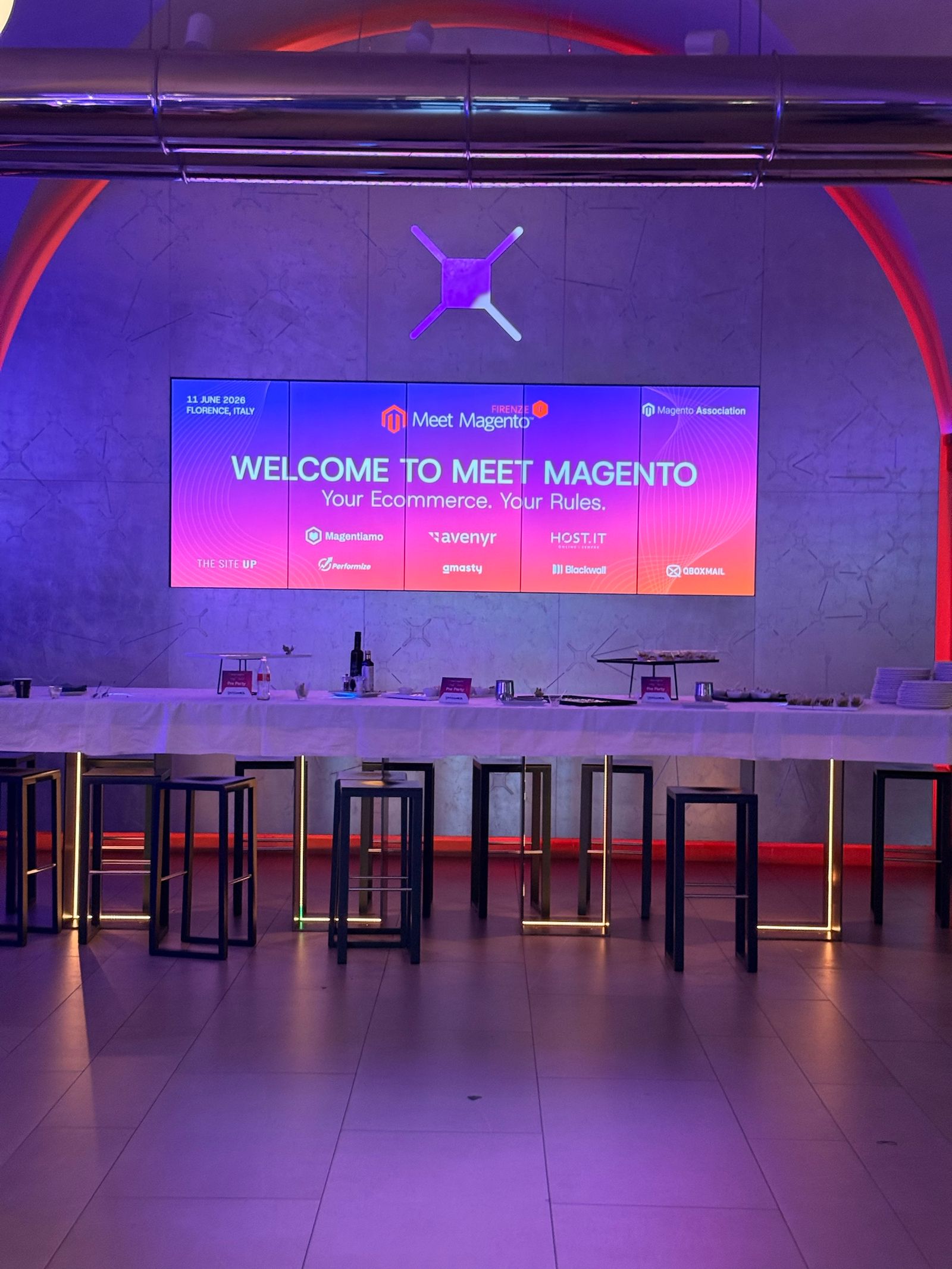 The main hall at Nana Bianca on the evening of 10 June 2026, lit in purple and red, with “Welcome to Meet Magento” on the screen ahead of the conference day.