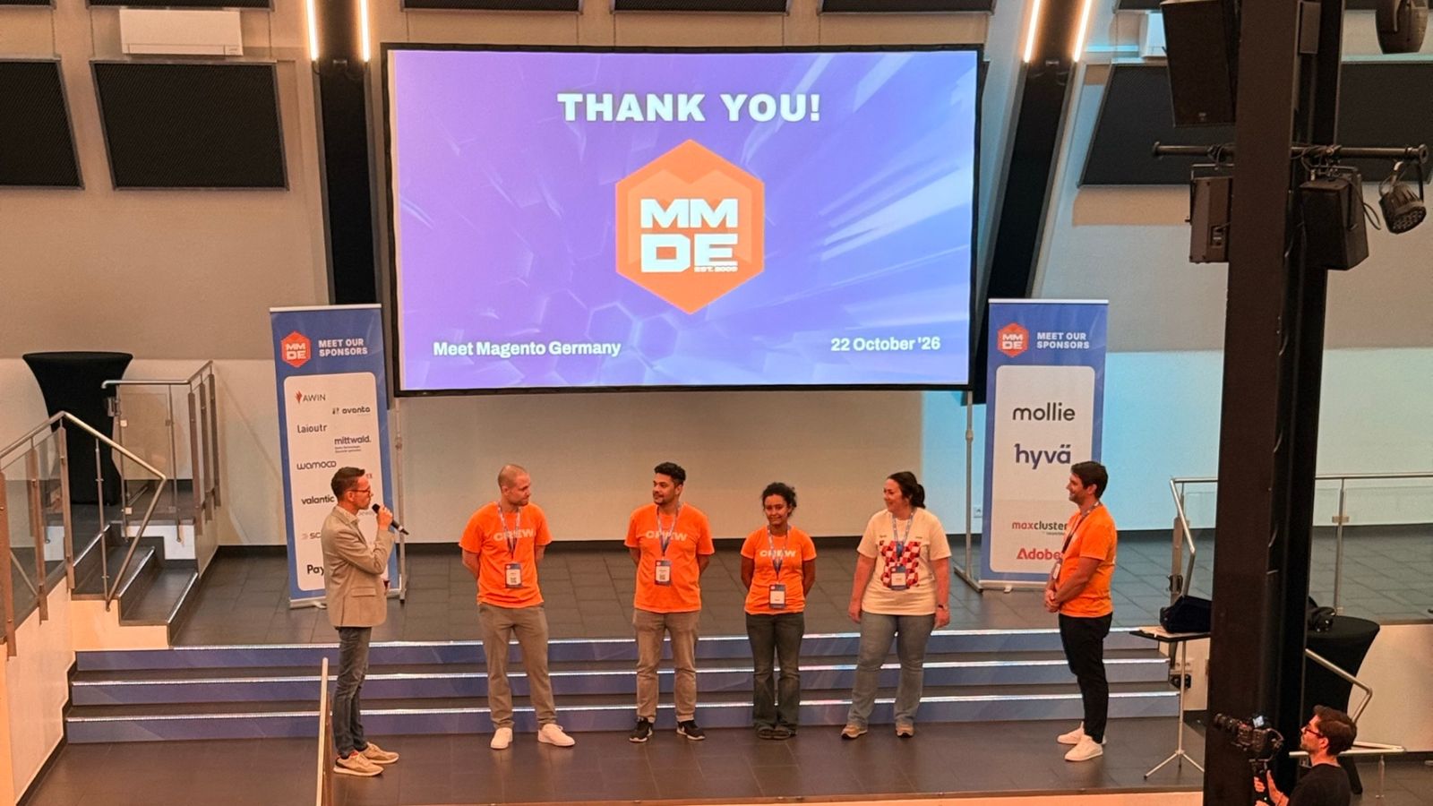 The Meet Magento Germany 2026 organisers and their crew in orange T-shirts on the main stage for the closing thank-you, in front of the hall.