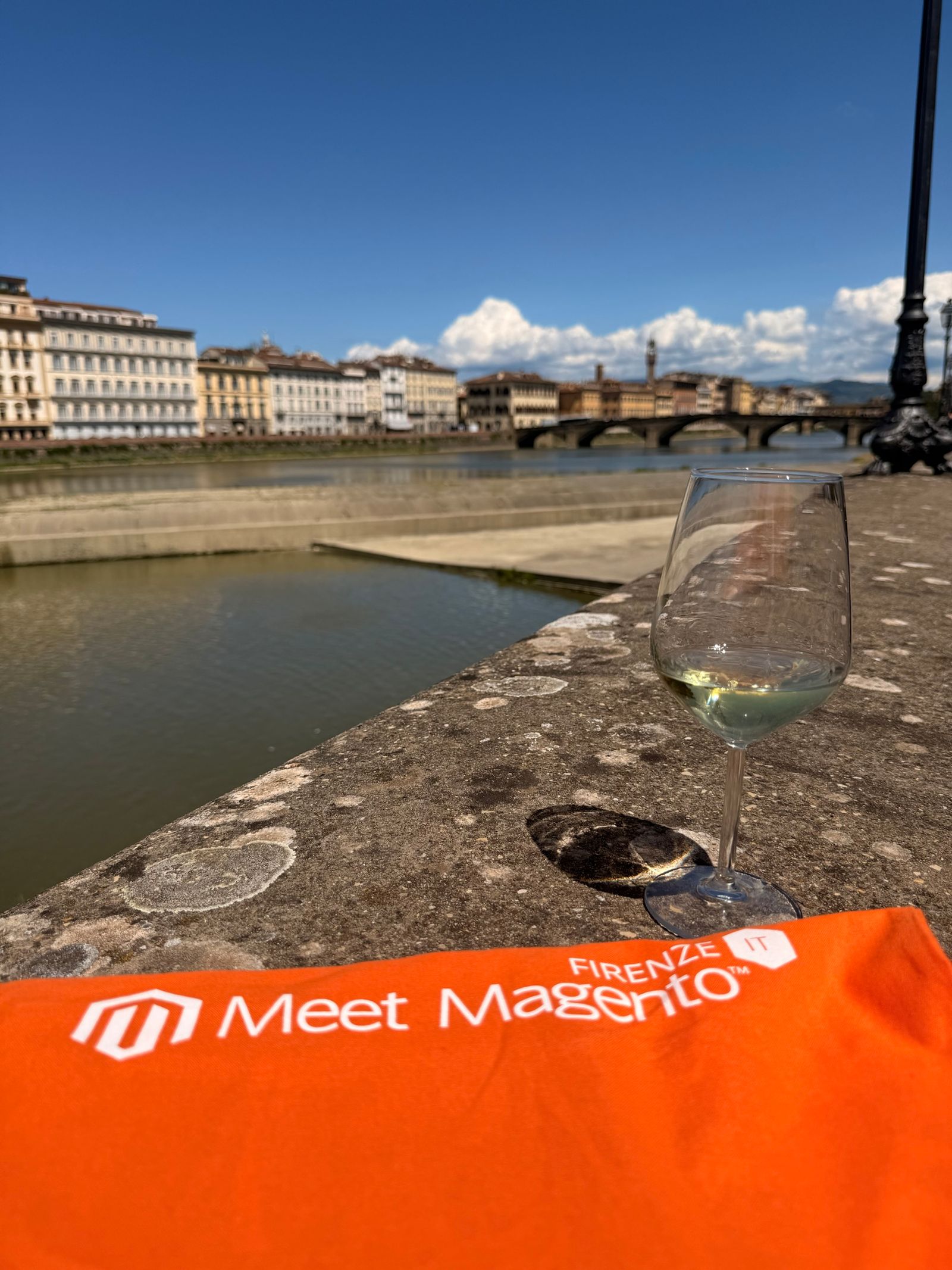 The orange Meet Magento Firenze banner and a glass of white wine on the stone wall of the Lungarno, with the Arno, a bridge and the Florence skyline behind them.