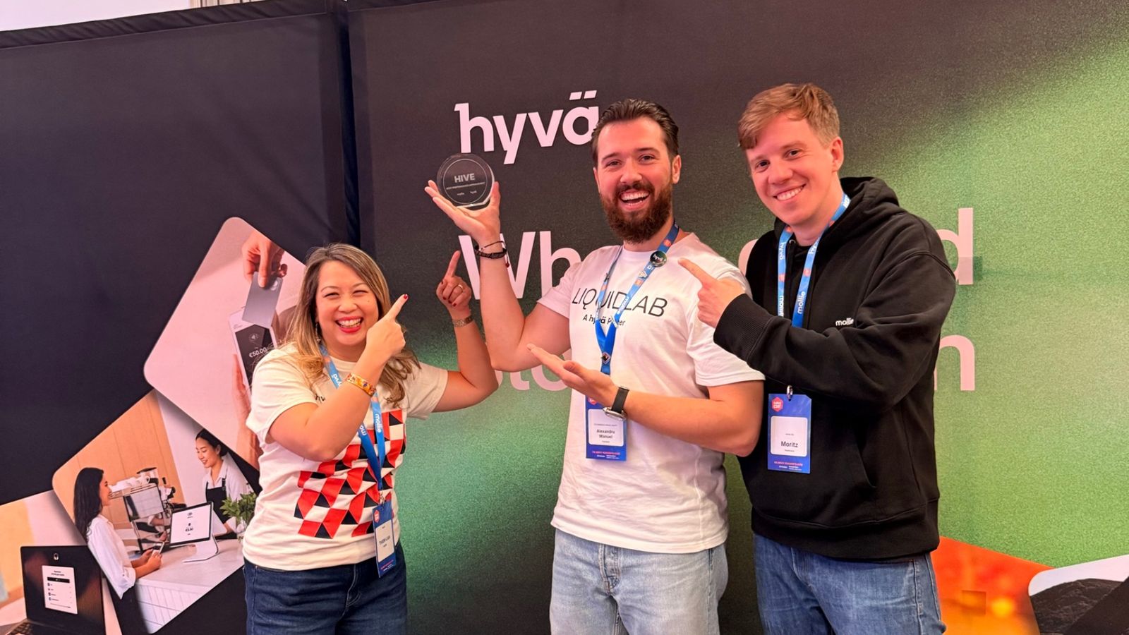 Thien-Lan de la Hyvä, Alexandru-Manuel Cărăbuș de la LIQUIDLAB și Moritz de la Mollie, la standul comun Hyvä și Mollie. Alexandru-Manuel, în tricoul „LIQUIDLAB, A Hyvä Partner”, ține sus trofeul HIVE, iar Thien-Lan și Moritz arată spre el.