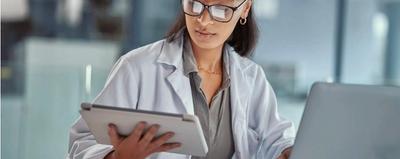 A doctor in a white coat reading a tablet beside an open laptop, with a stethoscope and paper records on the desk
