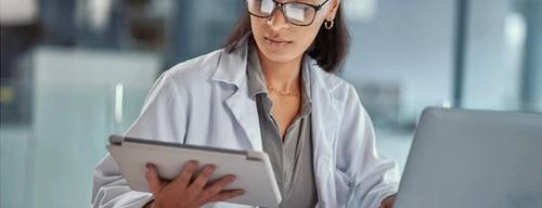 A doctor in a white coat reading a tablet beside an open laptop, with a stethoscope and paper records on the desk