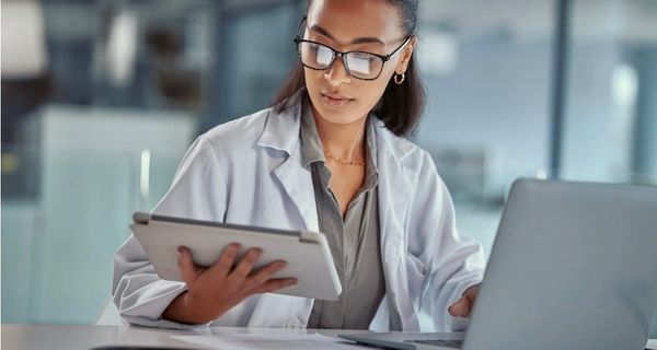 A doctor in a white coat reading a tablet beside an open laptop, with a stethoscope and paper records on the desk