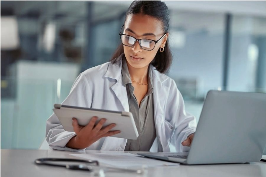 A doctor in a white coat reading a tablet beside an open laptop, with a stethoscope and paper records on the desk