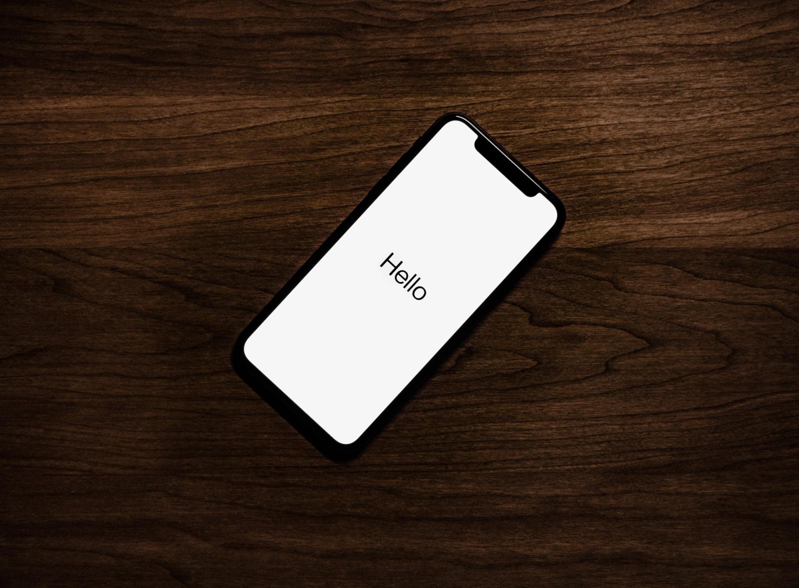 An iPhone laying on a dark wooden surface, with a white screen that says "Hello" in large black text