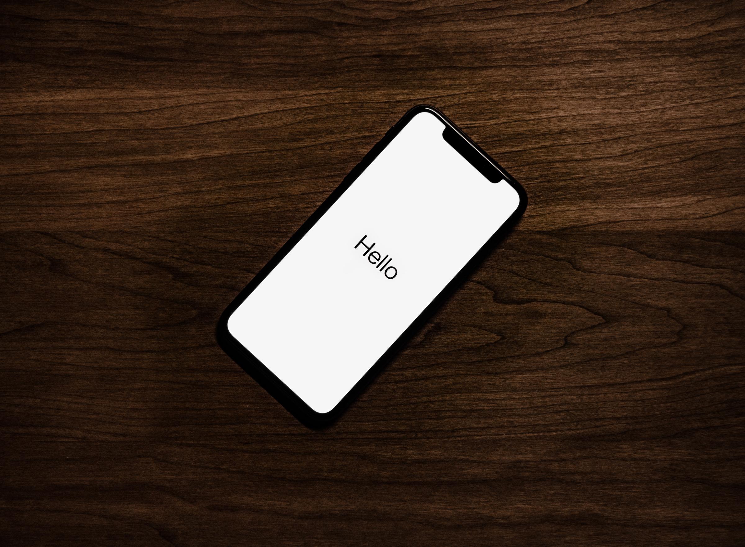 An iPhone laying on a dark wooden surface, with a white screen that says "Hello" in large black text