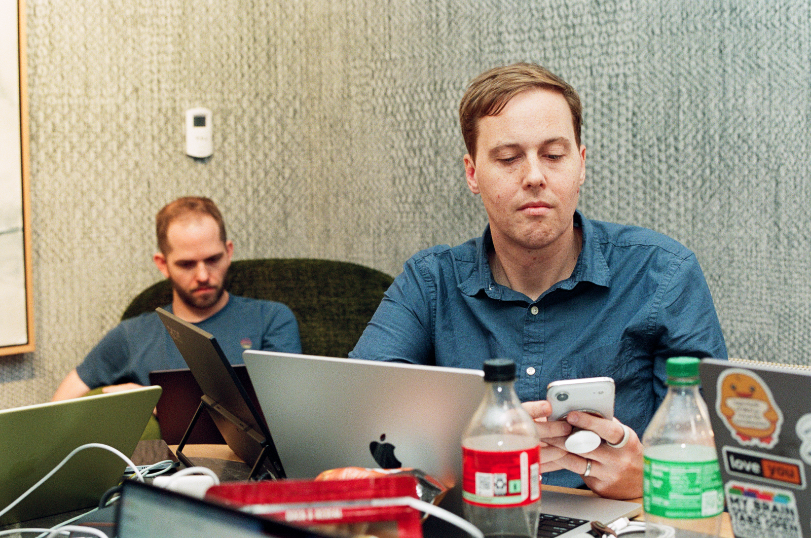Two Lickability engineers sitting near each other at a conference table, surrounded by MacBook laptops, soda bottles, and external monitors.