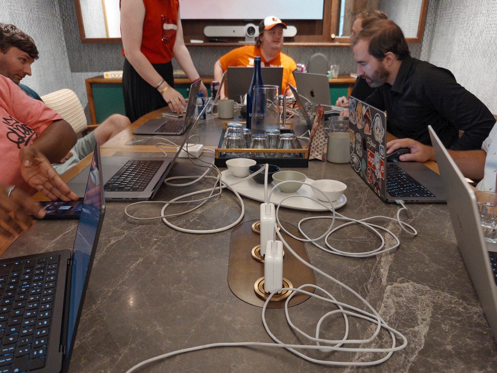 A conference table full of MacBook laptops with the center charging station occupied entirely by white charging bricks.