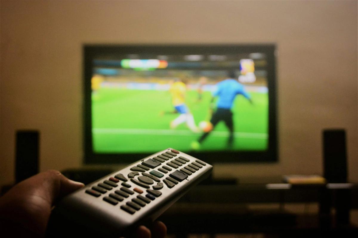 A hand holding a TV remote while watching football on television in a dark room, illustrating late nights and sleep debt