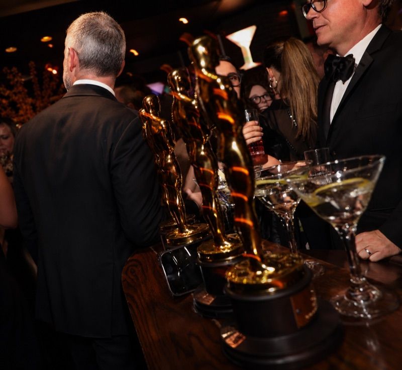 A row of Oscar statuettes on the dark wood bar beside martini glasses at an awards-night party