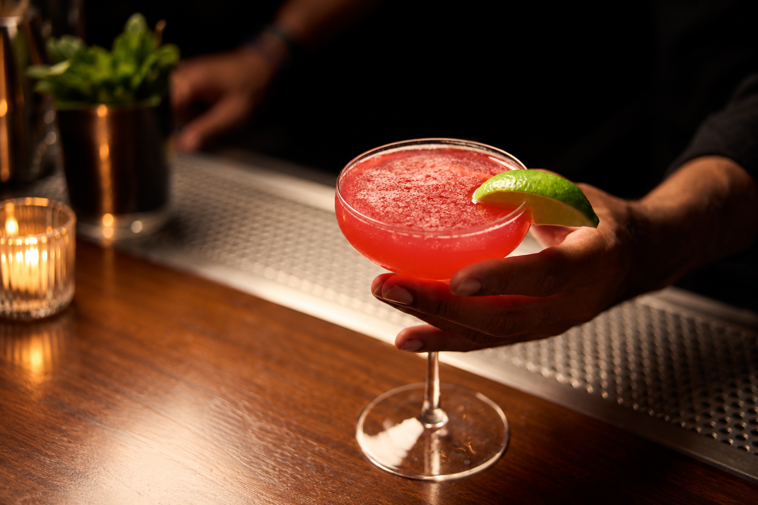 Bartender holding a bright red cocktail garnished with a lime wedge over a warmly lit wood bar.