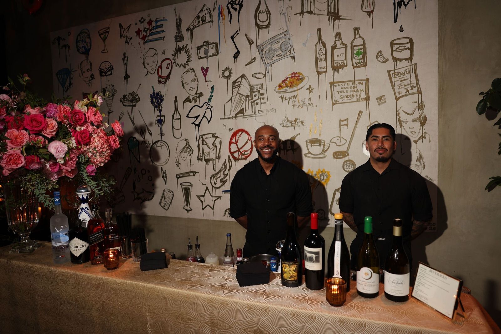 Two bartenders at a rose-decked bar in front of the large illustrated mural