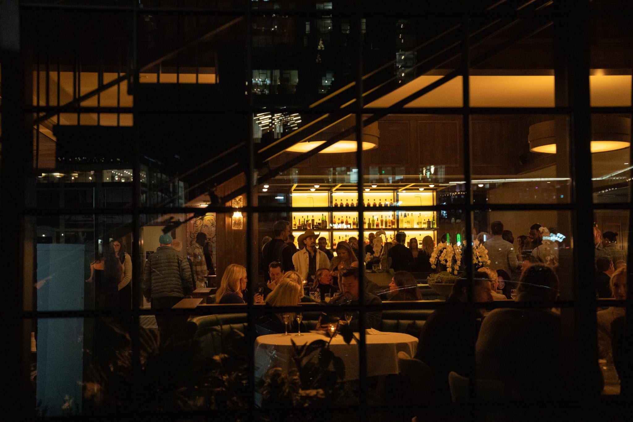 Evening view through steel-framed windows into the glowing dining room and bar at Craig's Nashville
