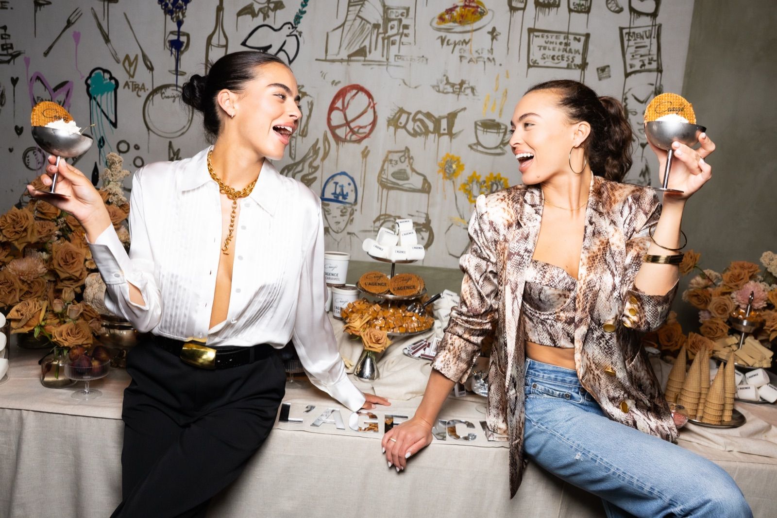 Two guests laughing over ice cream sundaes at a dessert table in front of the mural