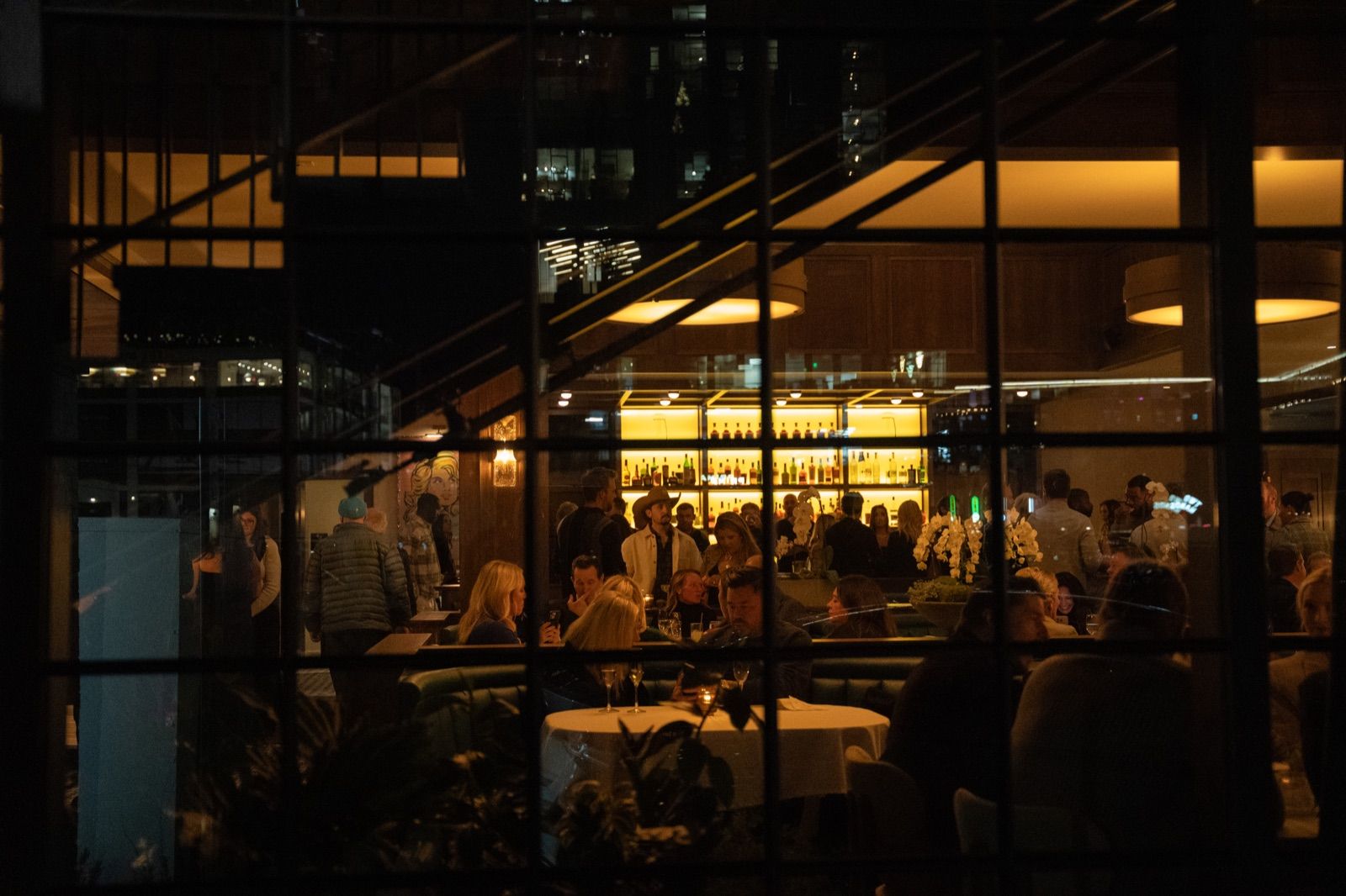Lit dining room and glowing backbar seen through steel-framed windows at night