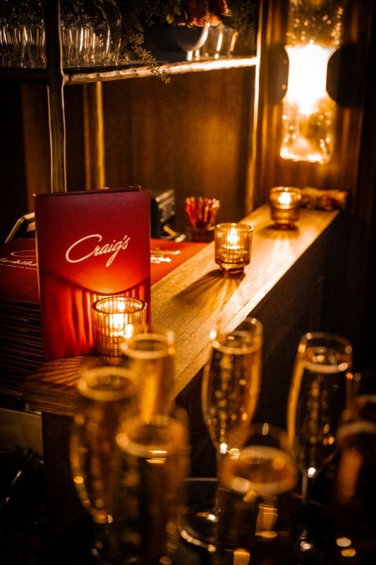 Craig's menu booklet and champagne flutes beside tea candles on a dimly lit bar shelf