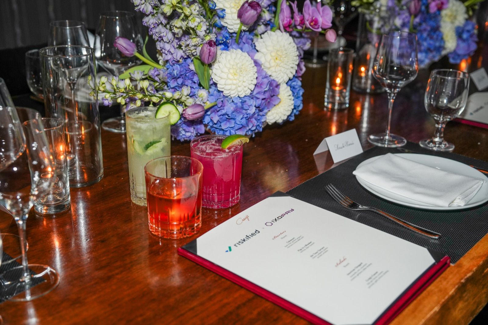 A place setting with colorful cocktails, a purple-and-white floral centerpiece, and an event menu card