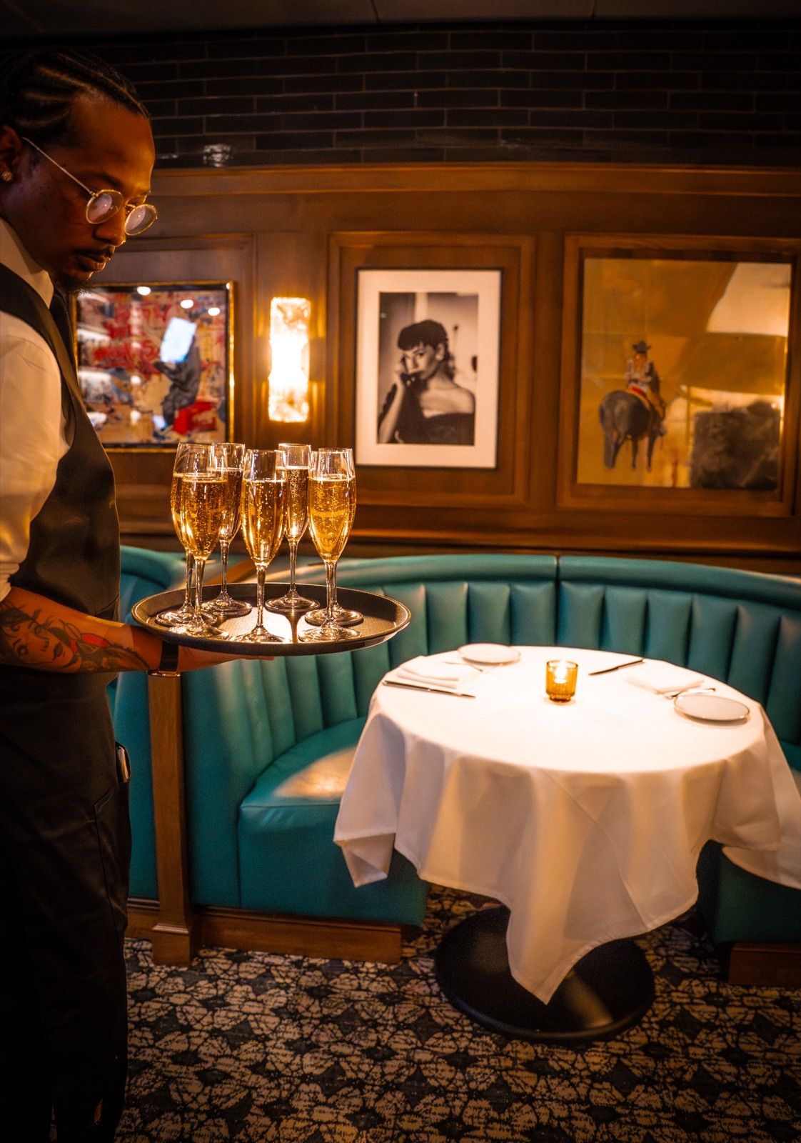 A server carrying a tray of six champagne flutes past a teal curved banquette