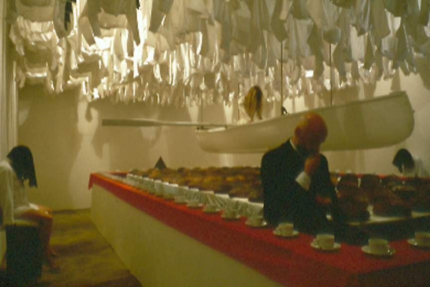 Michael Timpson, Who Blows Death’s Feathers: a dim interior with a long red table and a figure seated at the far end.